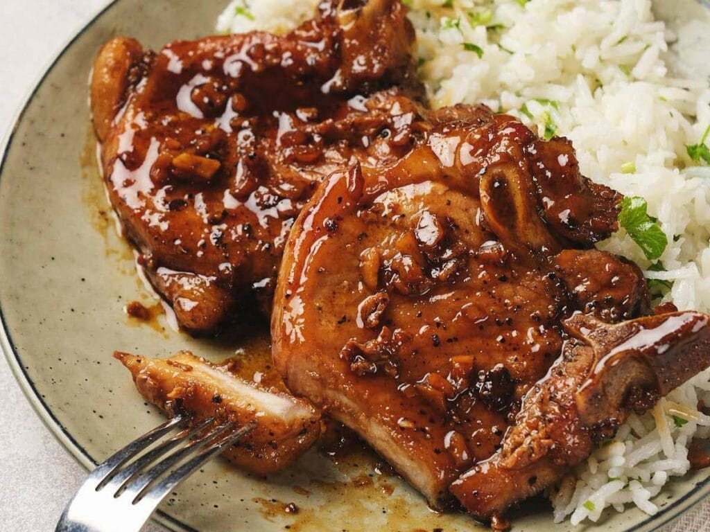 A plate with glazed grilled pork chops and white rice. A fork is positioned near the food.