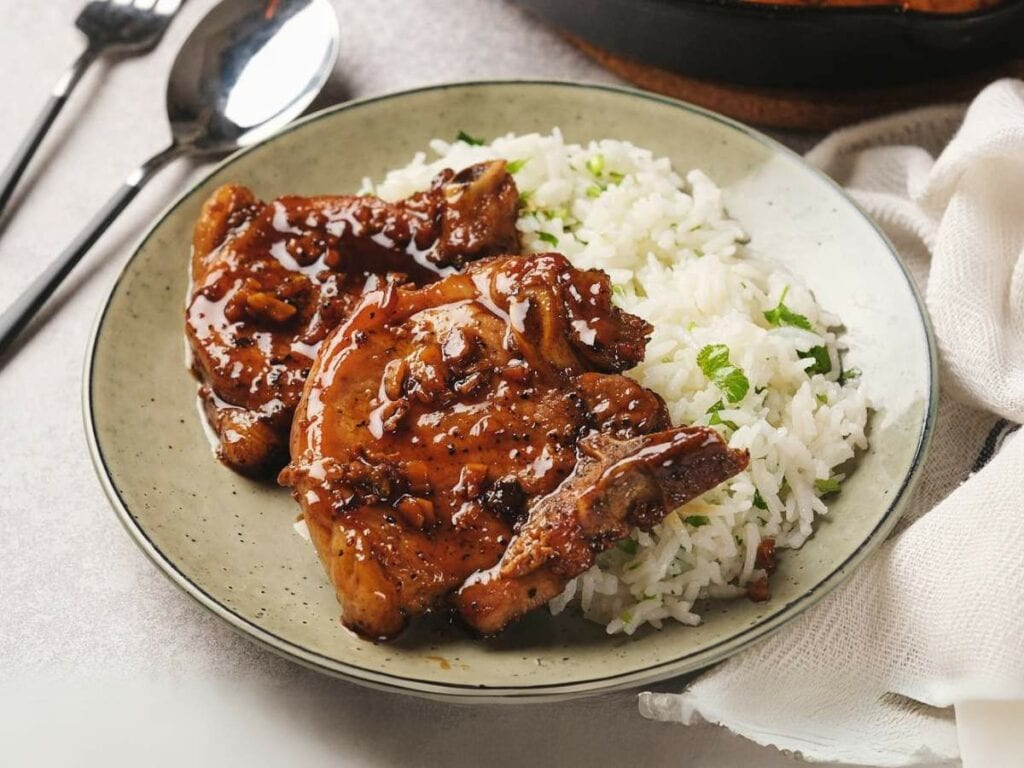 A plate with glazed pork chops and white rice garnished with herbs, accompanied by a fork and spoon on the side.