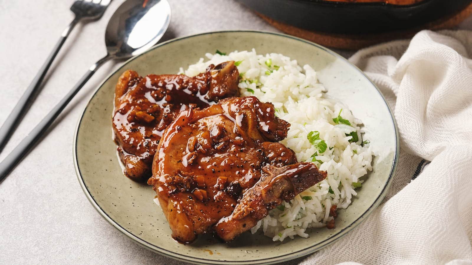 A plate with honey garlic pork chops glazed with sauce, accompanied by a serving of rice garnished with herbs. A fork and spoon are placed beside the plate.