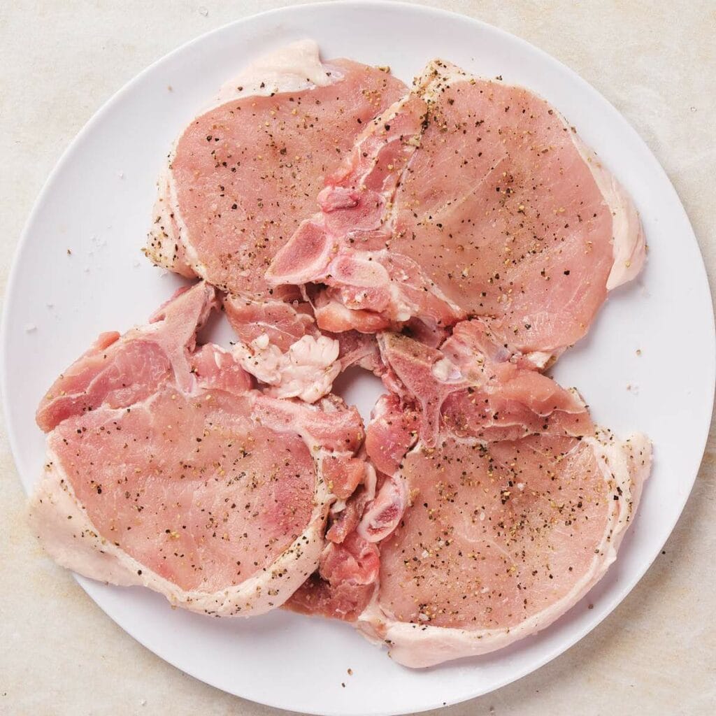 Four raw pork chops seasoned with pepper on a white plate.