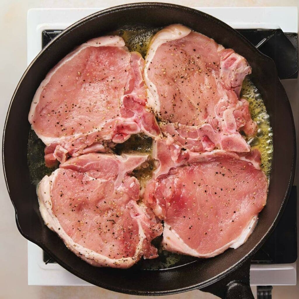 Four raw pork chops being cooked in a black frying pan on a stovetop, seasoned with pepper and sizzling in oil.