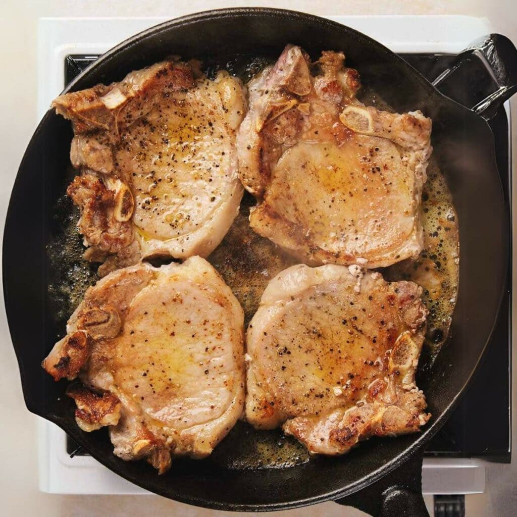 Four pork chops are being seared in a cast iron skillet on a stove. They are seasoned with salt and pepper.