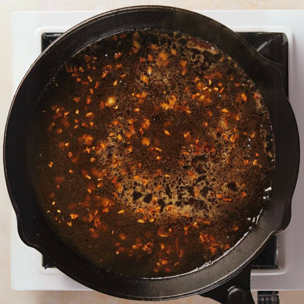 A cast iron skillet on a stove contains a simmering dark sauce with visible minced garlic and other small ingredients.