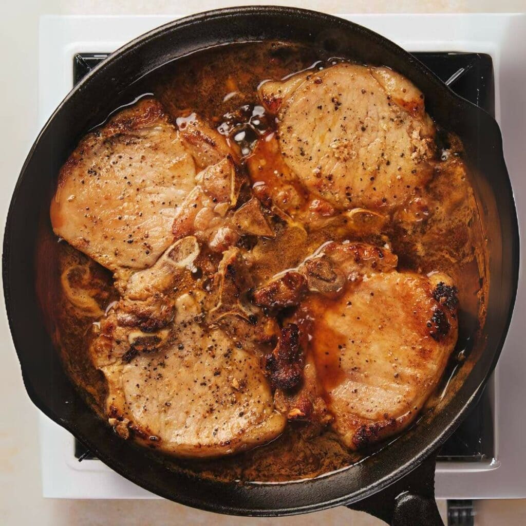 Four pork chops cooking in a skillet on a stovetop with a brown sauce and seasonings.