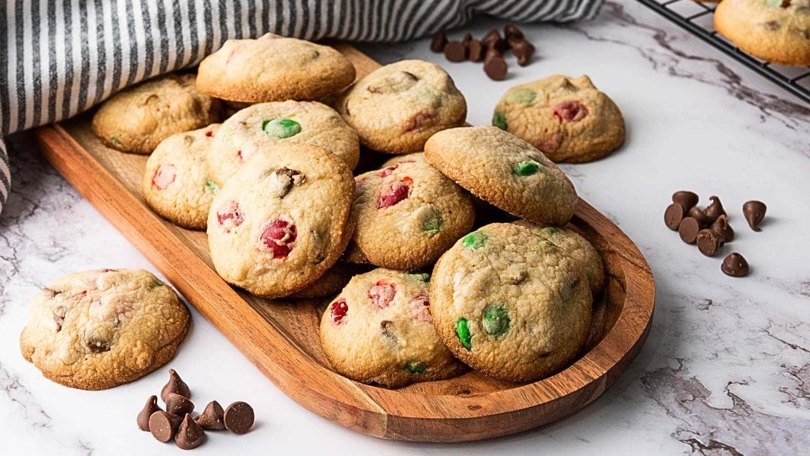 A wooden tray with freshly baked m&m christmas cookies containing chocolate chips and colored candies.