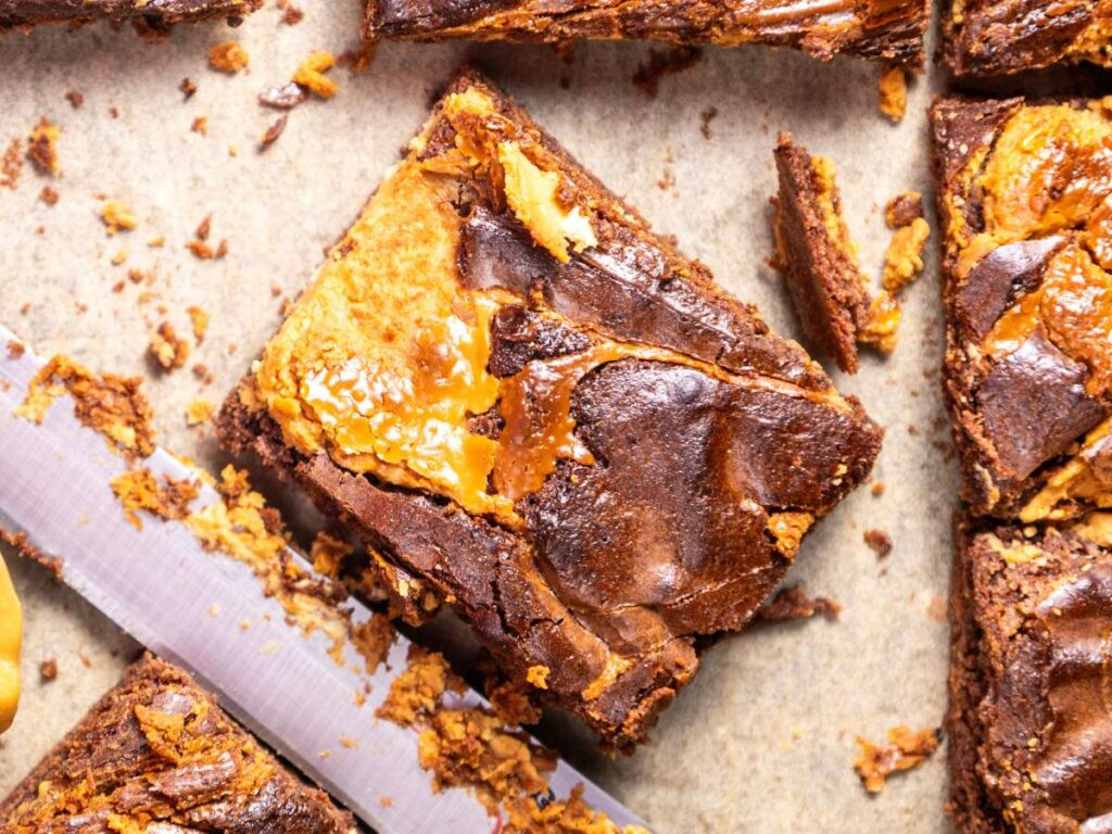 Peanut butter brownie squares with a caramel swirl on parchment paper, accompanied by a knife.