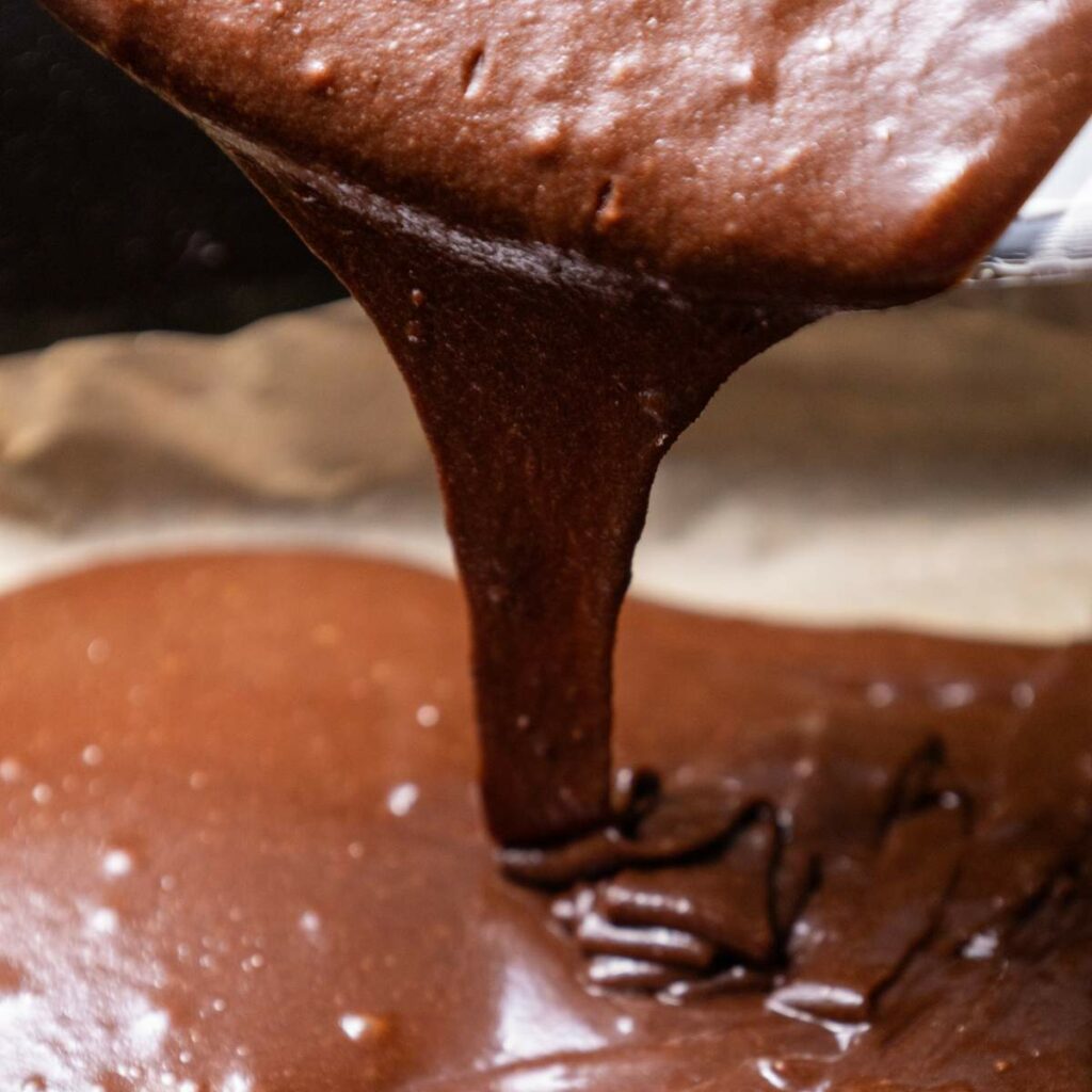 Close-up of chocolate batter being poured from a bowl into a baking pan lined with parchment paper.