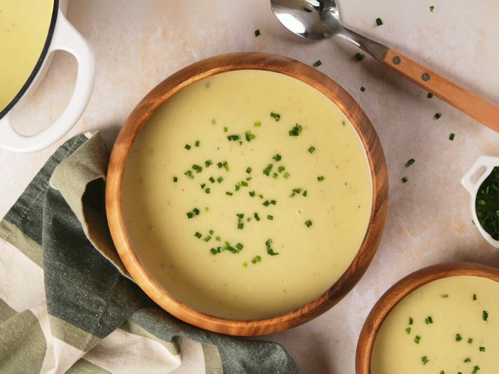 A wooden bowl of potato and leek soup garnished with chopped chives, accompanied by a spoon.