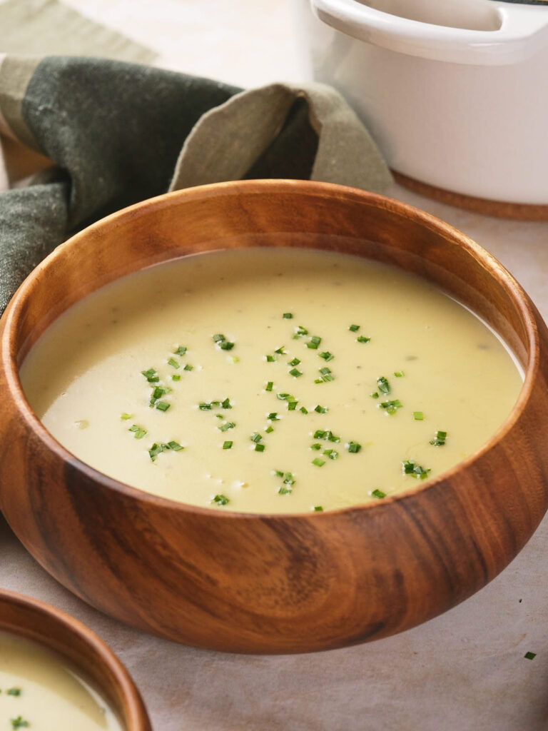 Wooden bowl of potato and leek soup garnished with chopped chives, placed on a light surface.