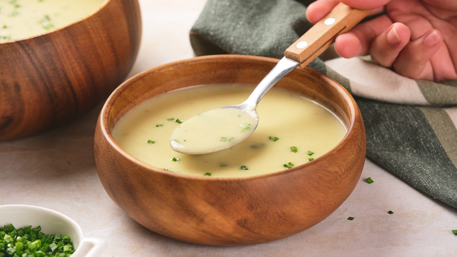 A person holds a spoon of poato and leek soup with chives over a wooden bowl, with another bowl and a striped napkin nearby.