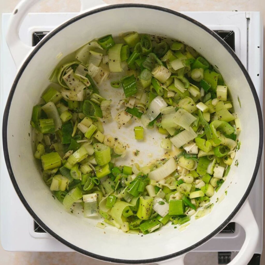 Chopped green onions and leeks sautéing in a white pot on a stovetop.