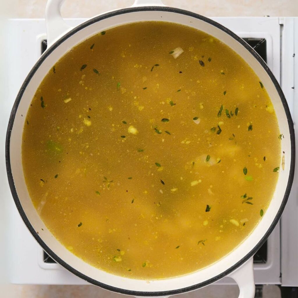 A pot of soup with herbs and diced vegetables is simmering on a stovetop.