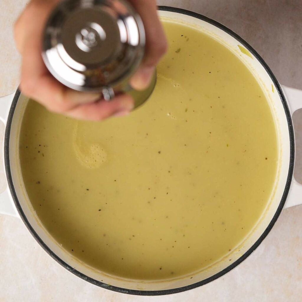 Hand seasoning a pot of creamy green soup with a shaker.