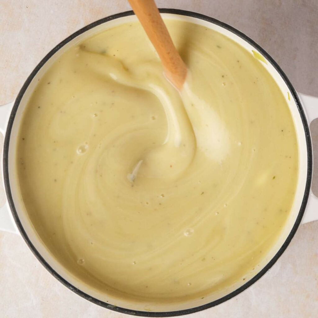 A pot filled with creamy yellow soup being stirred by a wooden spoon.