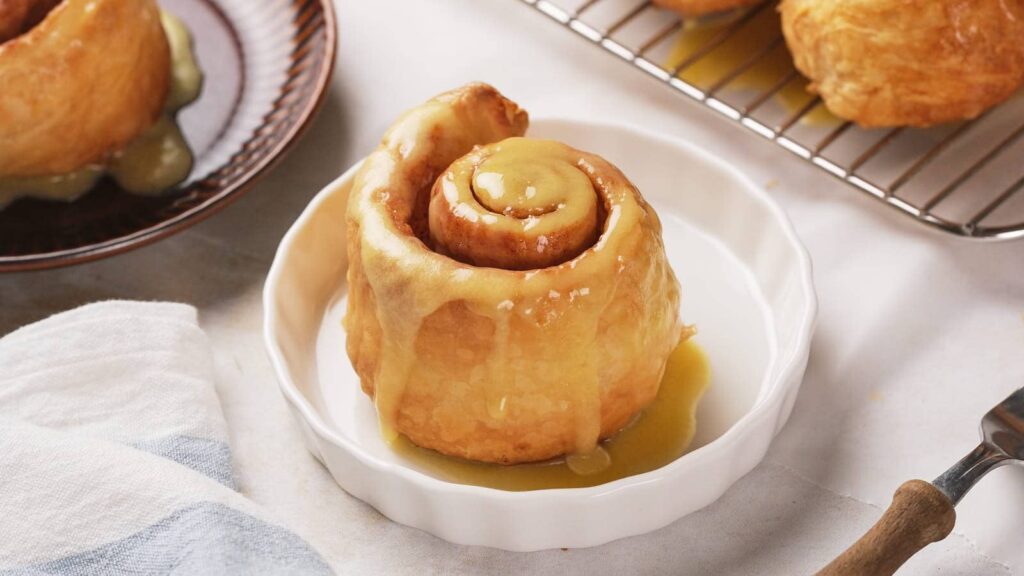 Puff pastry cinnamon roll sits on a white plate, with more rolls on a wire rack in the background.