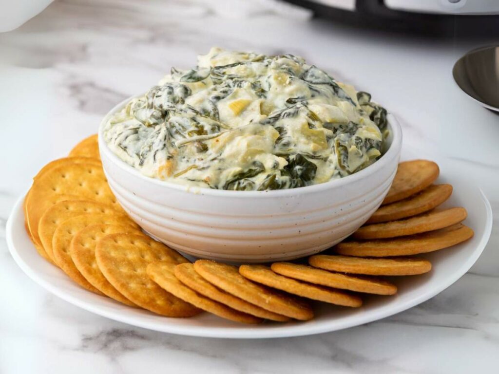 A bowl of creamy spinach dip surrounded by a ring of round crackers on a white plate, set on a marble surface.