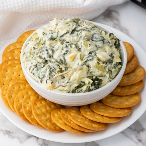 A bowl of spinach artichoke dip surrounded by round crackers on a white plate, placed on a marble countertop.