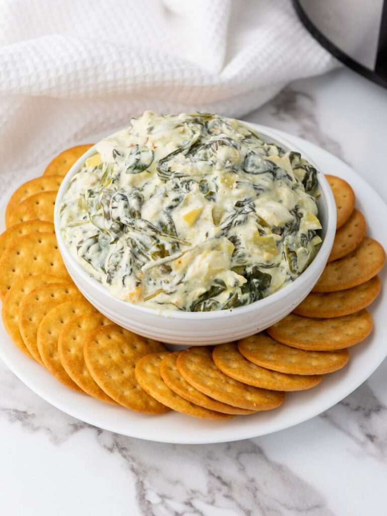 A bowl of spinach artichoke dip surrounded by round crackers on a white plate, placed on a marble countertop.