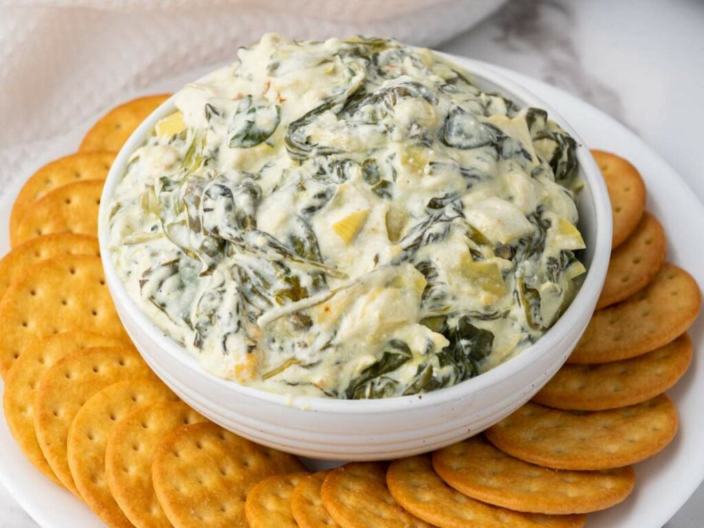 A bowl of creamy spinach artichoke dip is surrounded by round, golden crackers on a white plate.