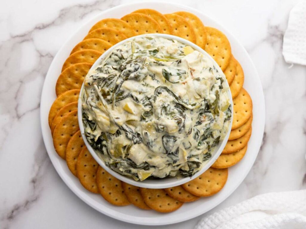 A bowl of spinach artichoke dip is centered on a plate, surrounded by round crackers, on a marble surface.