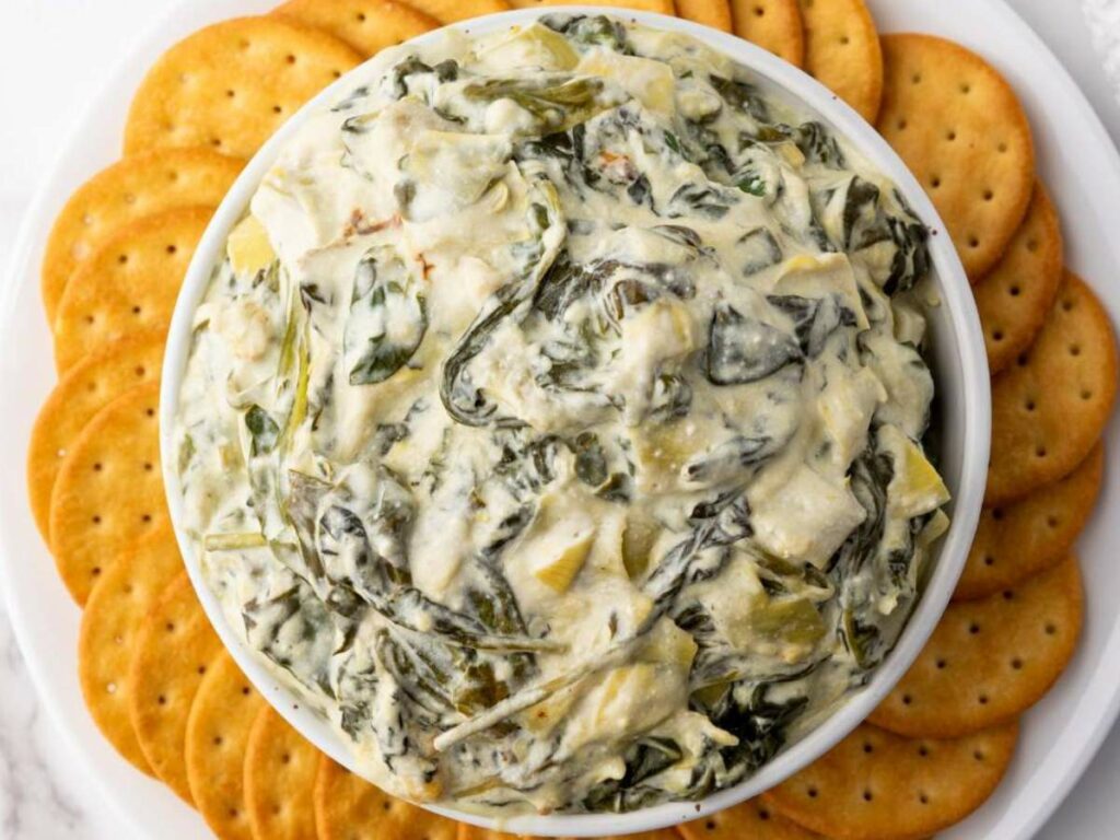 A bowl of spinach artichoke dip is centered on a plate surrounded by round crackers.