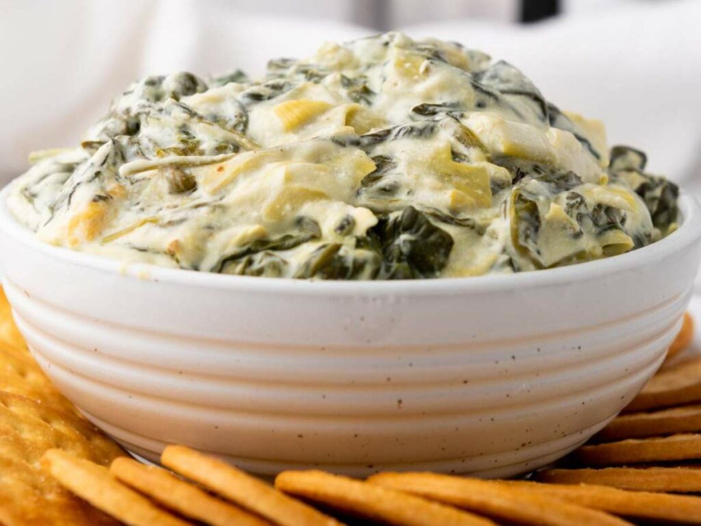 A white bowl filled with creamy spinach artichoke dip, surrounded by round crackers.