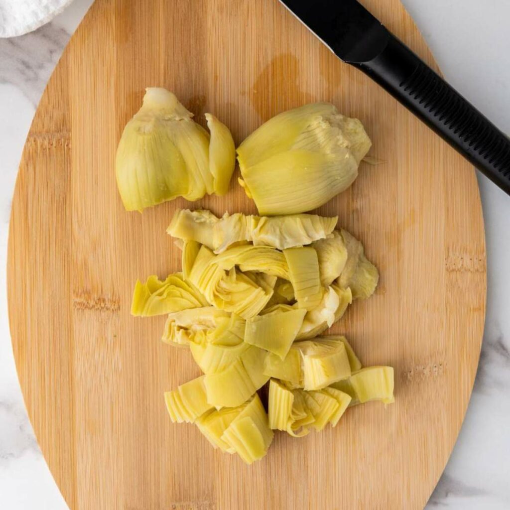 Chopped artichokes and a black peeler on a wooden cutting board.