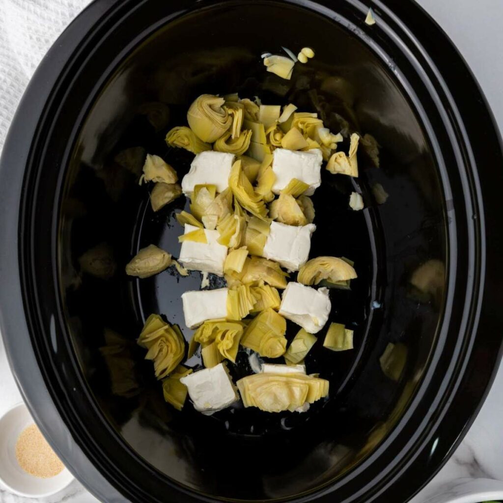 Chopped artichoke hearts and cubes of cream cheese in a black slow cooker.