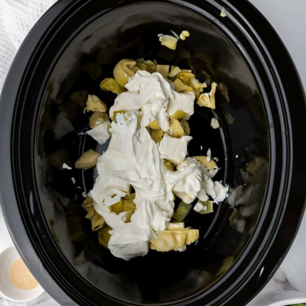 Artichokes and cream cheese in a black slow cooker, placed on a marble surface.