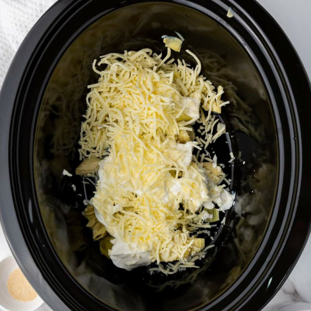 Cheese, sour cream, and artichokes in a black slow cooker, ready for cooking.