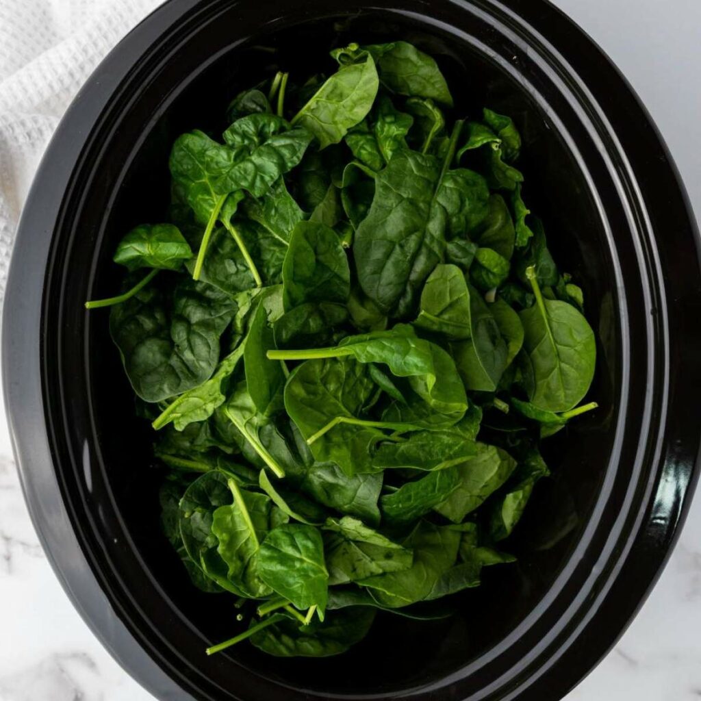 A black slow cooker filled with fresh spinach leaves on a marble surface.