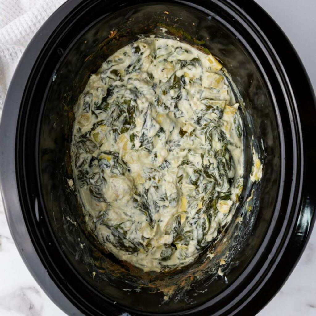 A slow cooker filled with creamy spinach artichoke dip.