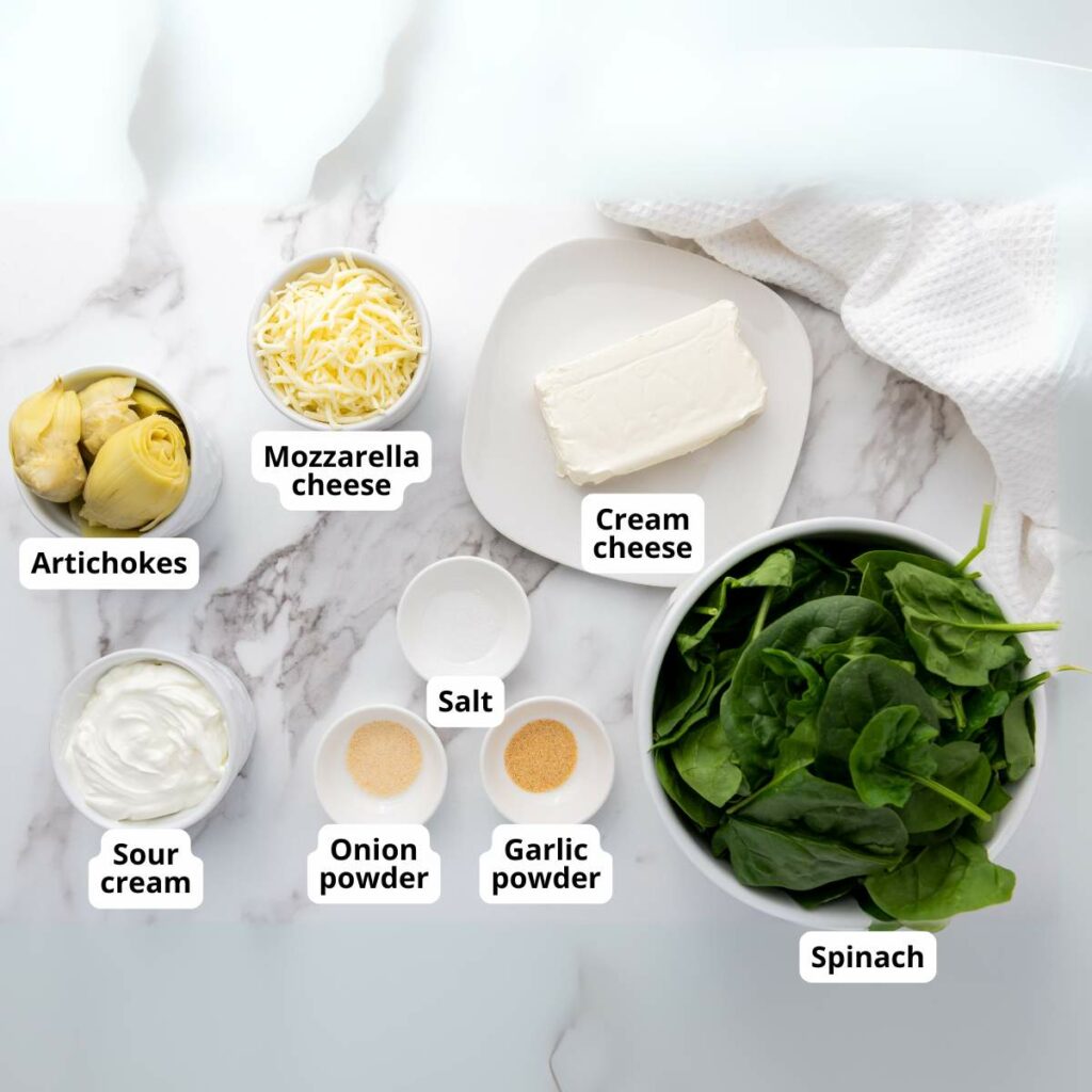 Ingredients for a spinach artichoke dip are arranged on a marble surface.