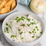 A bowl of creamy caramelized onion dip topped with chopped green onions and black pepper, next to a bowl of crackers. An onion and striped cloth are in the background.