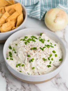 A bowl of creamy caramelized onion dip topped with chopped green onions and black pepper, next to a bowl of crackers. An onion and striped cloth are in the background.