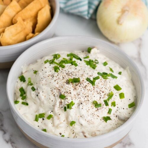 A bowl of creamy caramelized onion dip topped with chopped green onions and black pepper, next to a bowl of crackers. An onion and striped cloth are in the background.