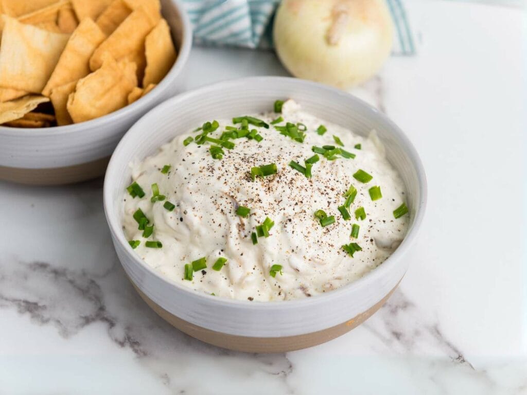 A bowl of creamy caramelized onion dip garnished with chopped chives and black pepper.