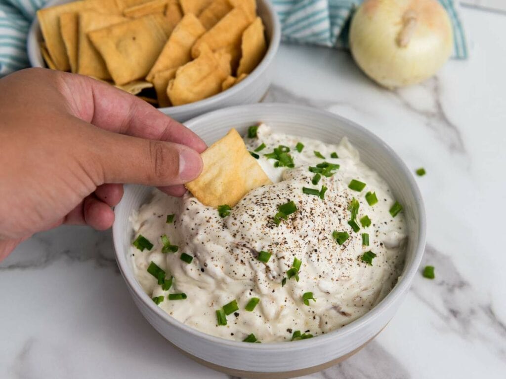 Hand dipping a caramelized onion chip into a bowl of creamy dip topped with chopped chives and pepper.