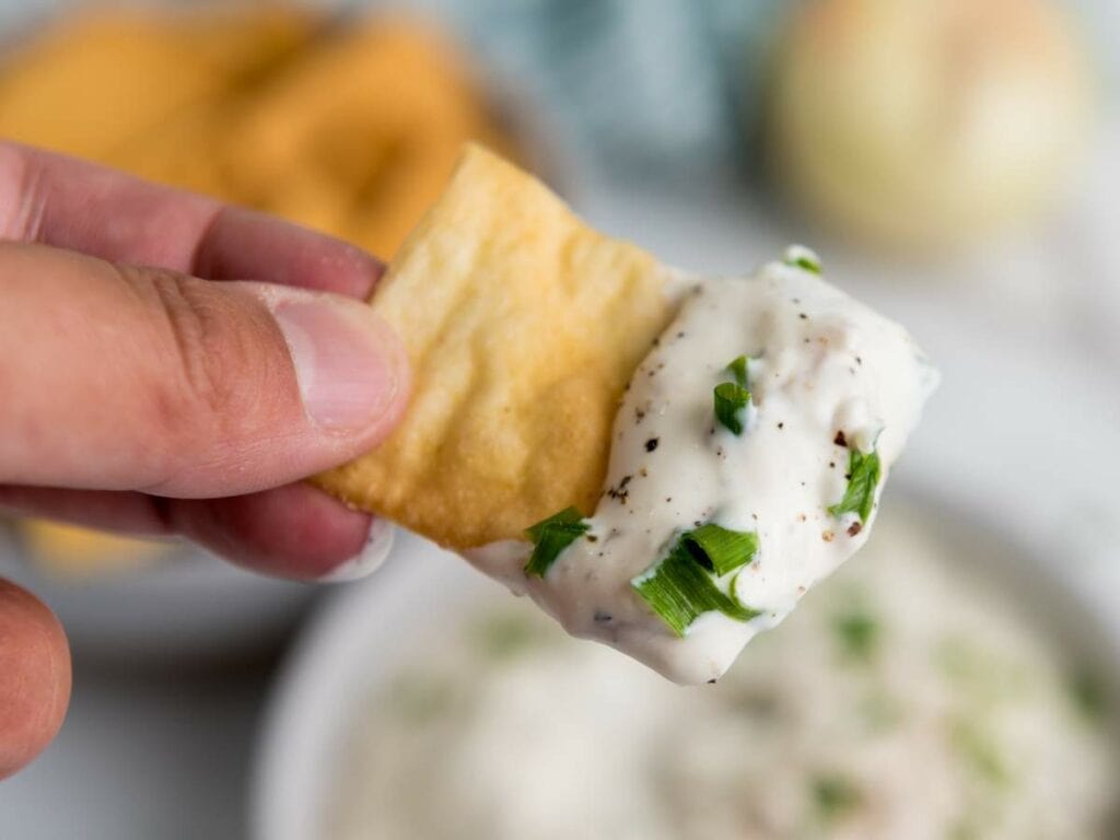 Close-up of a hand holding a cracker dipped in creamy, herb-flecked white sauce.