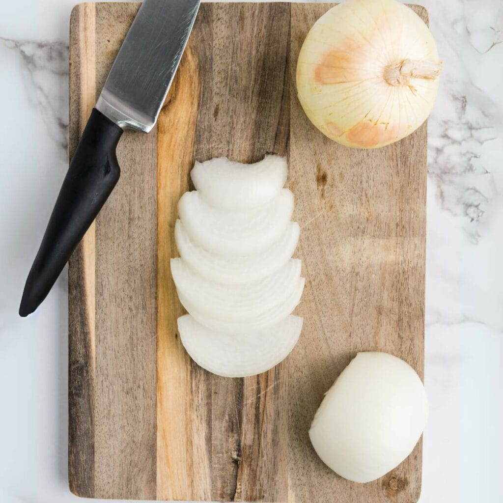 A knife and sliced onion are on a wooden cutting board, along with a whole and half onion.