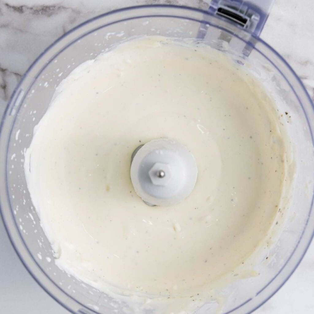 Top view of a food processor with smooth, creamy white sauce on a marble surface.