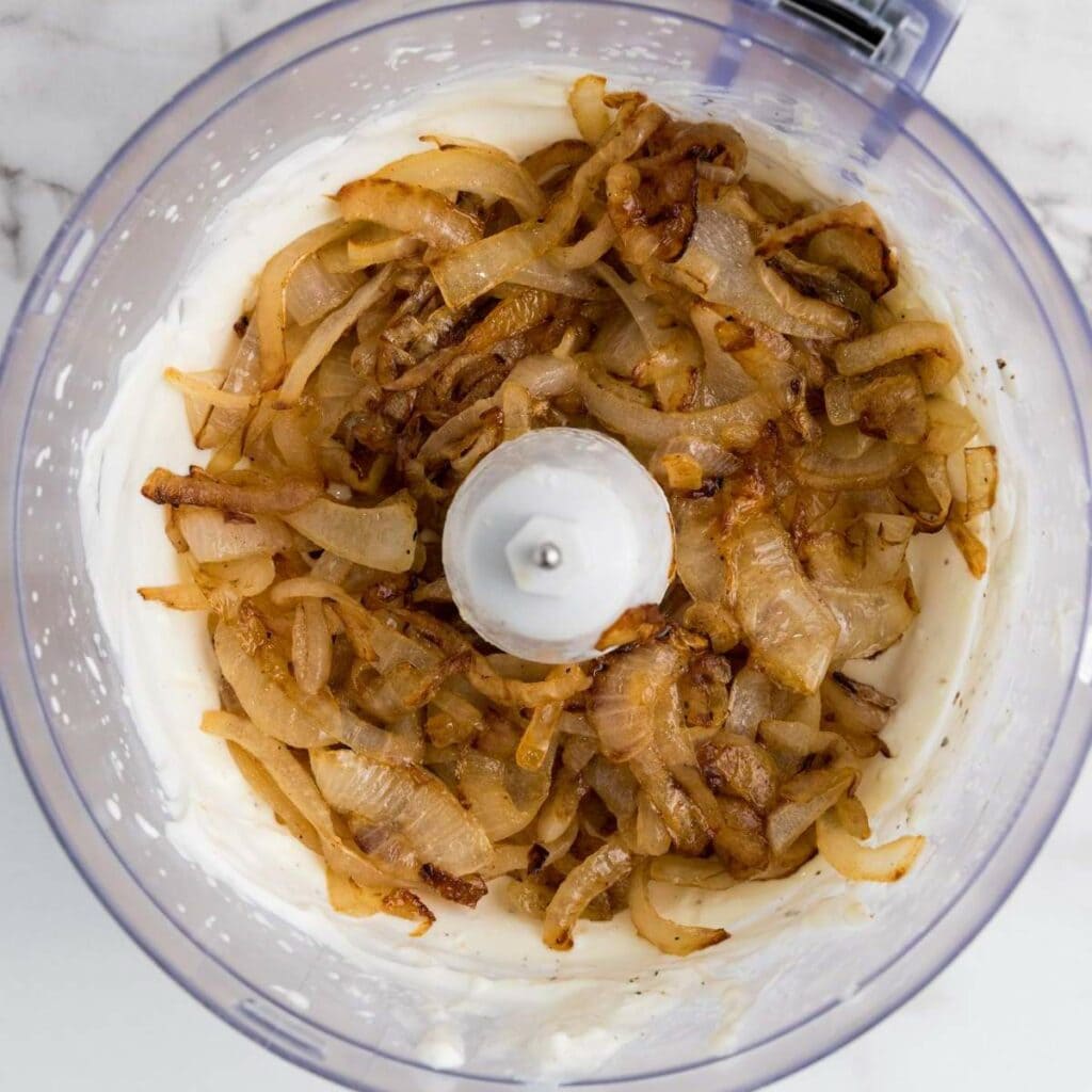 Caramelized onions in a food processor bowl on top of a creamy mixture, seen from above.