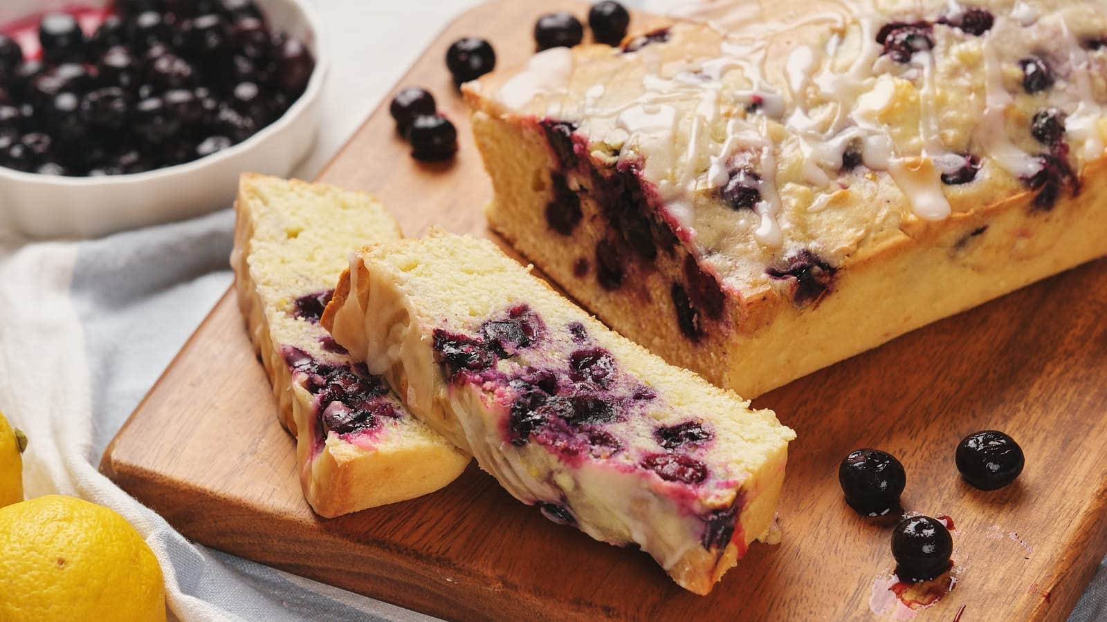 Lemon blueberry loaf with icing on a wooden board, accompanied by whole blueberries and lemons.