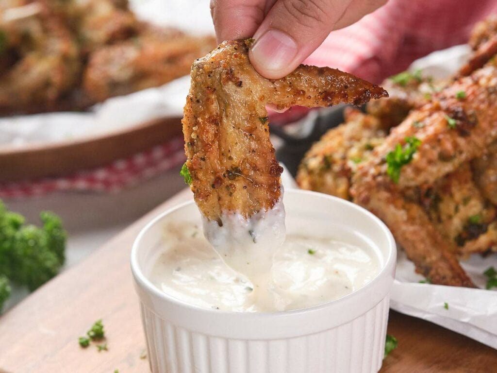 A hand dips a parmesan and garlic wings into a white sauce in a ramekin.