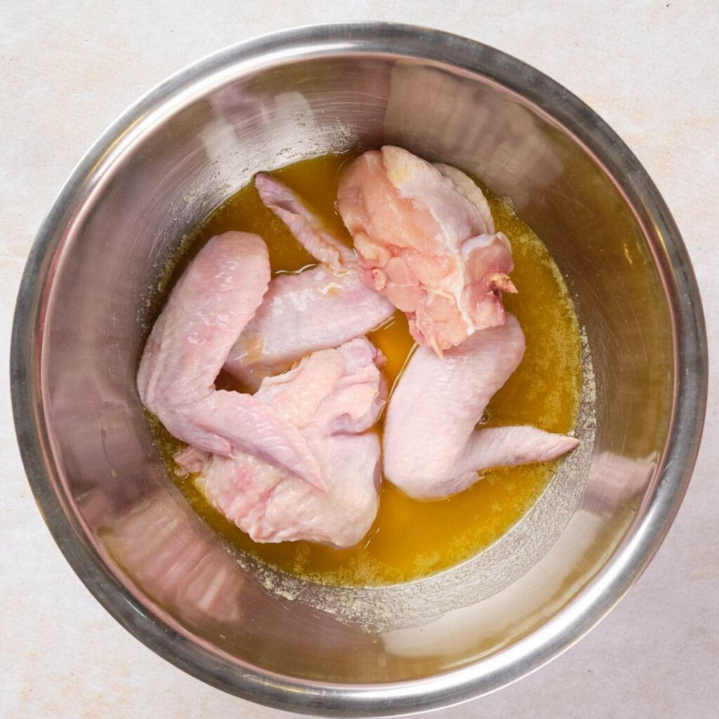 Raw chicken wings marinating in a metal bowl with a yellow liquid.