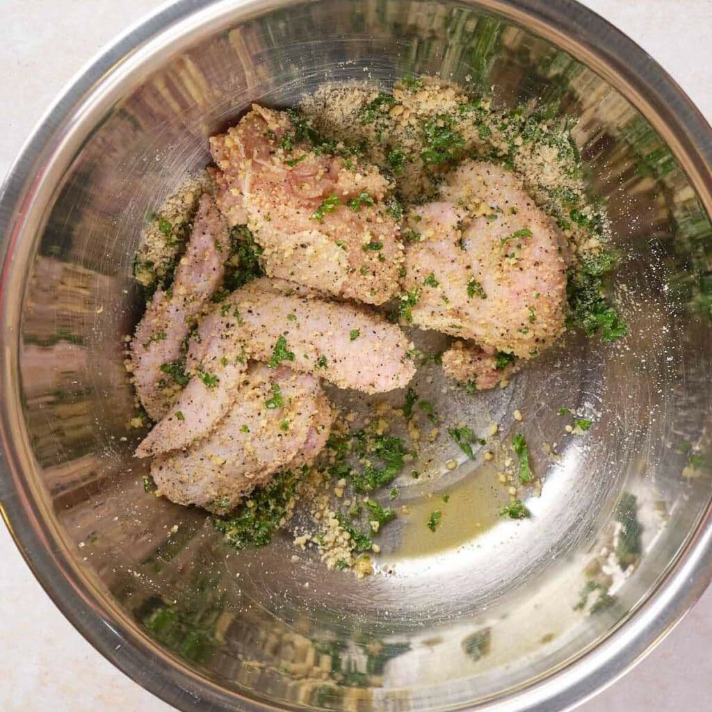 Raw chicken pieces marinated with herbs, spices, and breadcrumbs in a metal bowl.